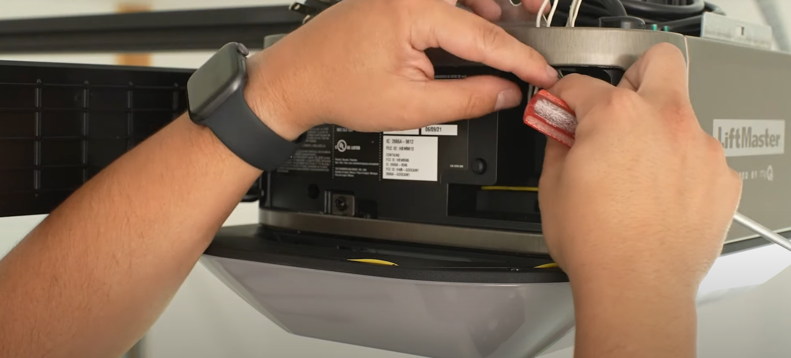 A technician in a blue uniform repairing a garage door opener motor unit mounted on the ceiling of a residential garage.